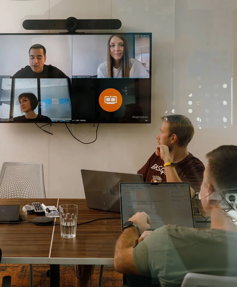 A couple of guys in a conference room facing the screen where an online video conference is ongoing