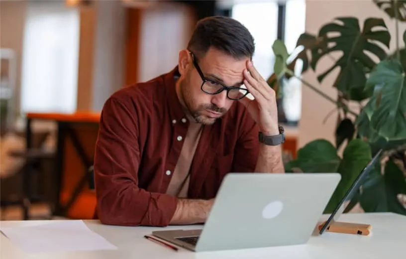 A man looking down on his laptop while a hand touches his forehead