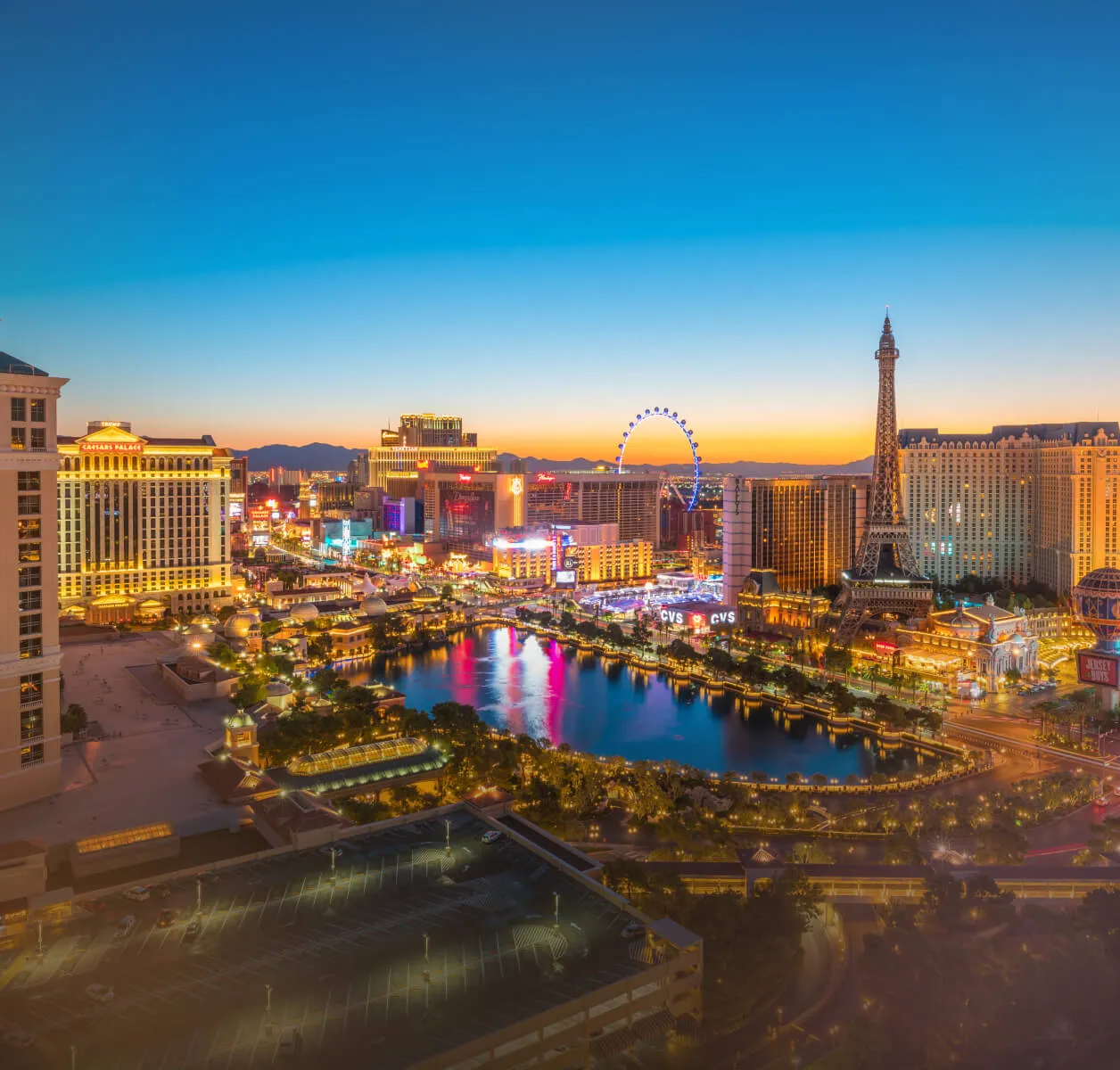 High-angle twilight view of the Las Vegas Strip featuring the Bellagio fountains, the Eiffel Tower at Paris Las Vegas, and the High Roller observation wheel under a sunset sky.