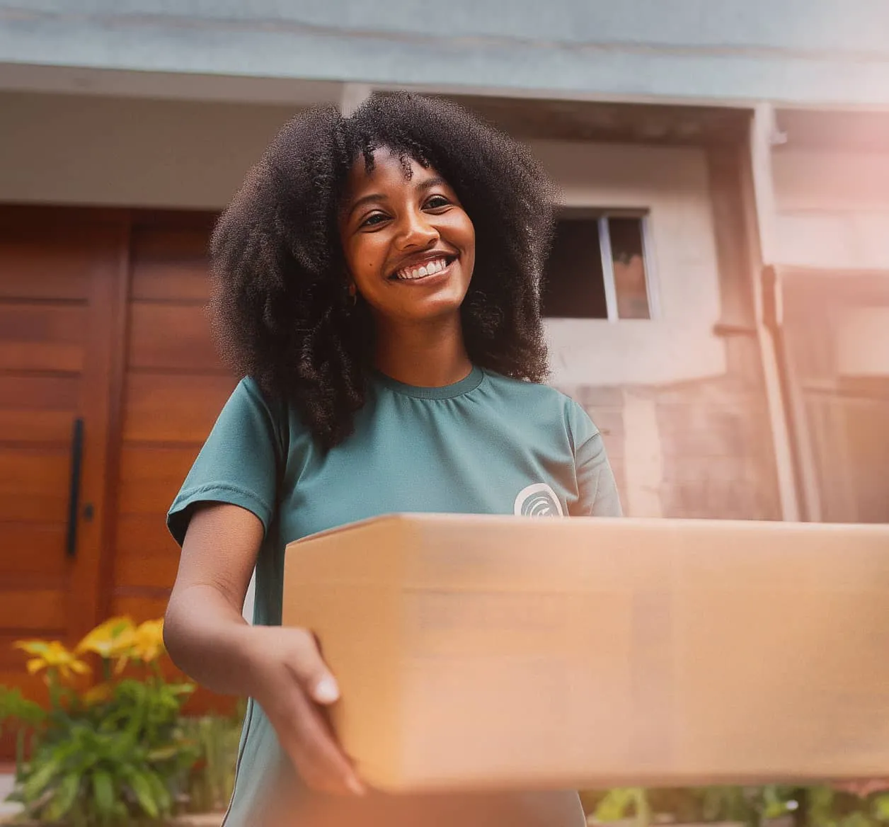 A volunteer standing outside of a home is handing someone a cardboard box of goods.