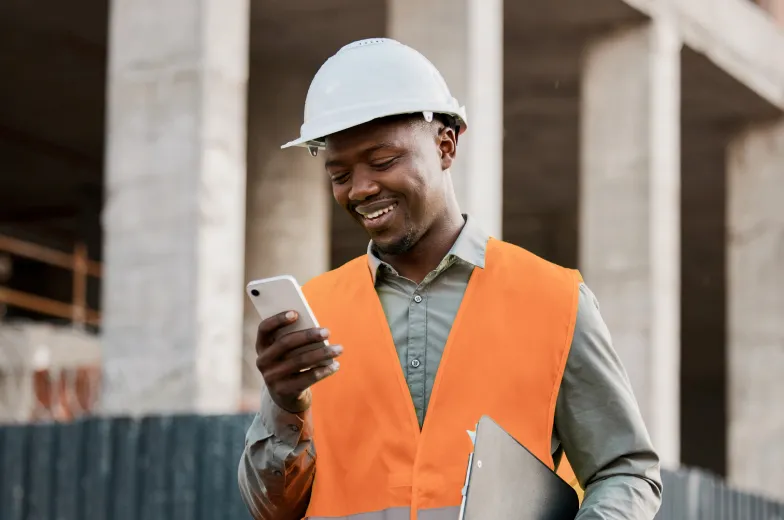 A man wearing a safety helmet and vest looks at his mobile phone