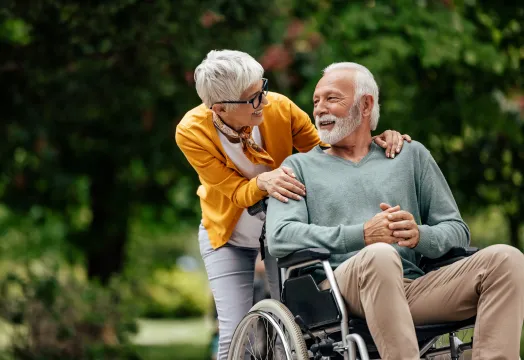 An elderly man in a wheelchair is with his wife, both appearing happy in a bright outdoor environment.