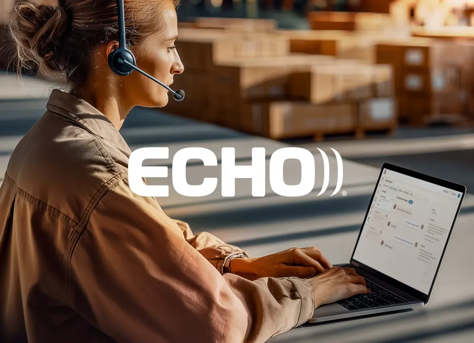 A woman in a headset works on a laptop inside a sunlit warehouse filled with stacked boxes.