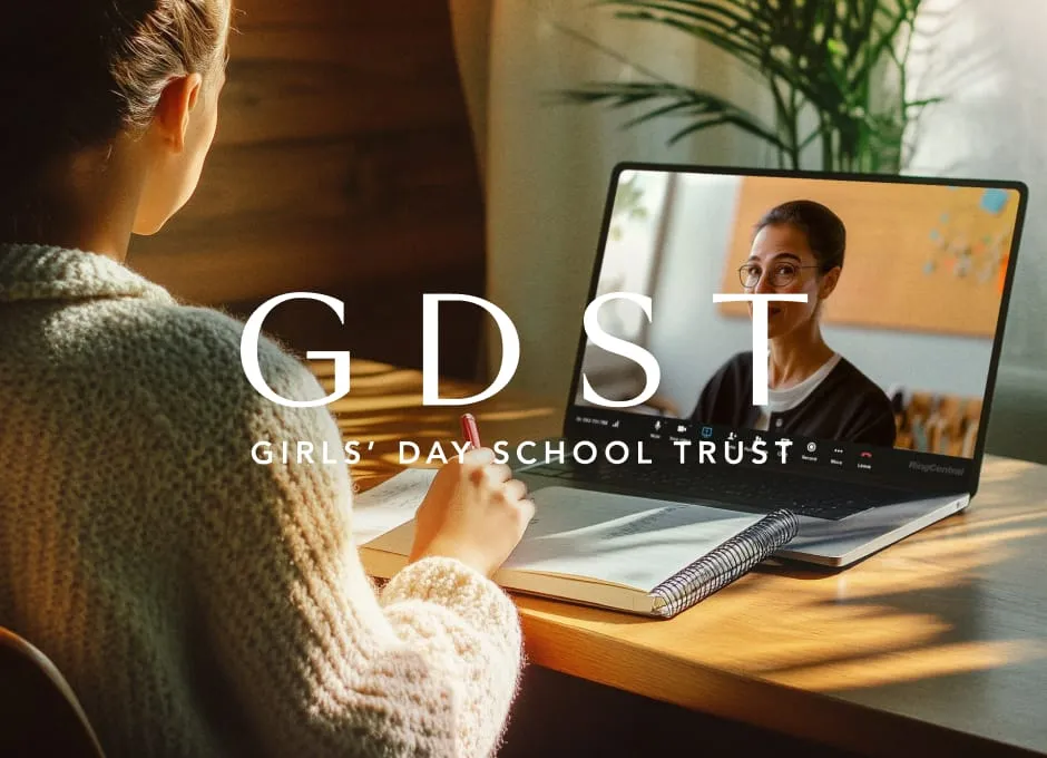 A student takes notes while participating in a video call with a teacher on a laptop in a sunlit room.